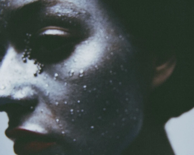 Close-up portrait of a person with silver body paint and water droplets, focusing on the face and side profile.