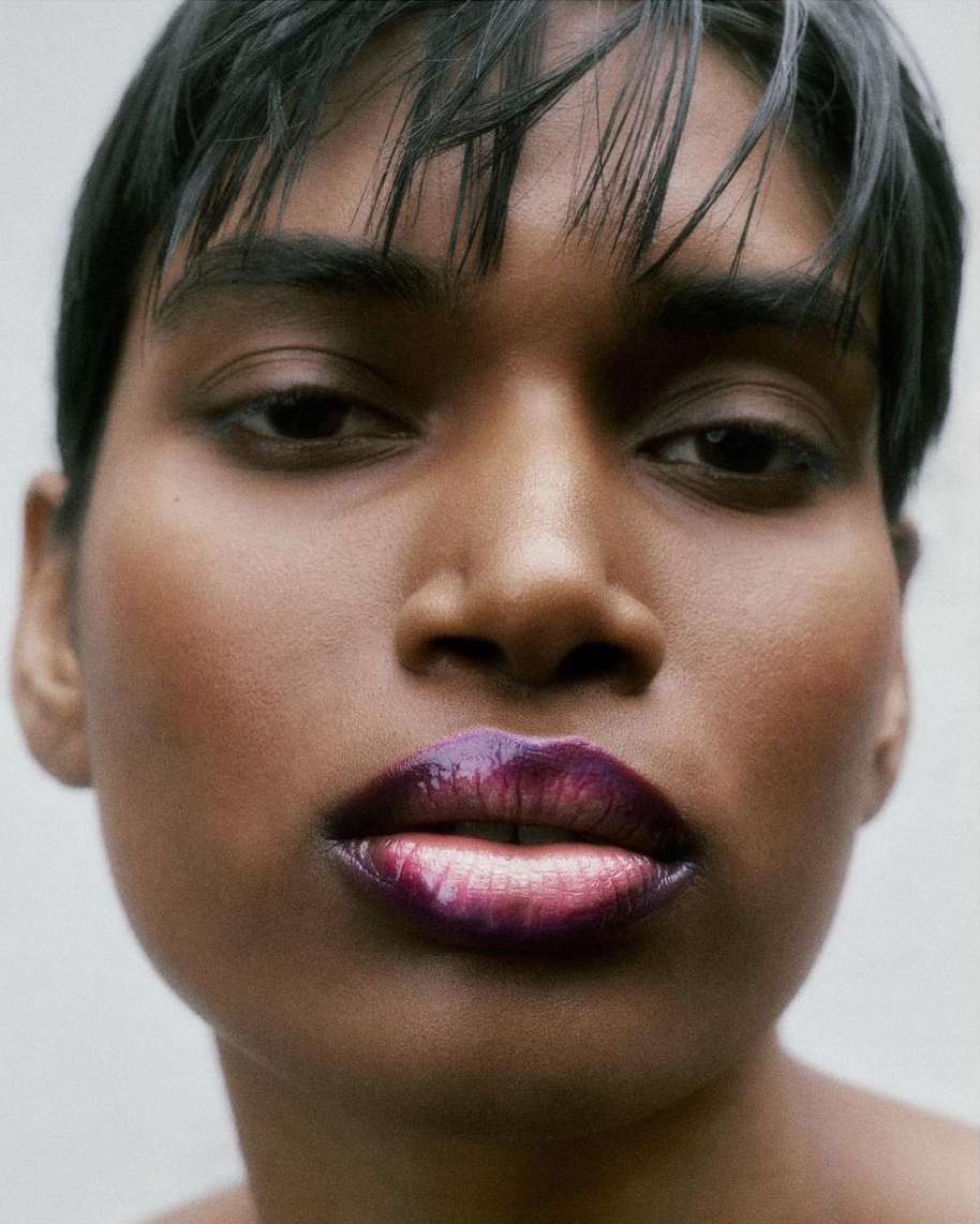 Close-up portrait of a young adult female with dark skin, short dark hair, and shiny ombre purple lipstick against a plain white background.