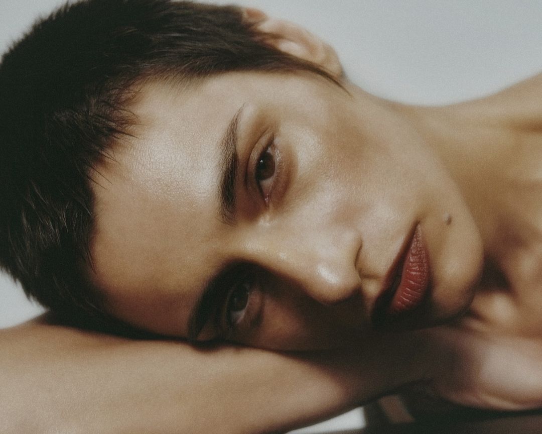 Close-up studio portrait of a woman with dark short hair, wearing red lipstick, resting her face on her arm, neutral expression.