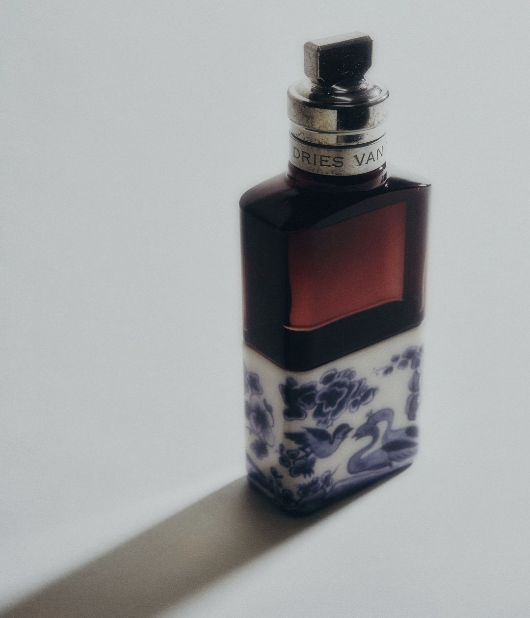 Cologne bottle by Dries Van Noten with red liquid in the upper section and white and blue floral design on the bottom, studio lit.