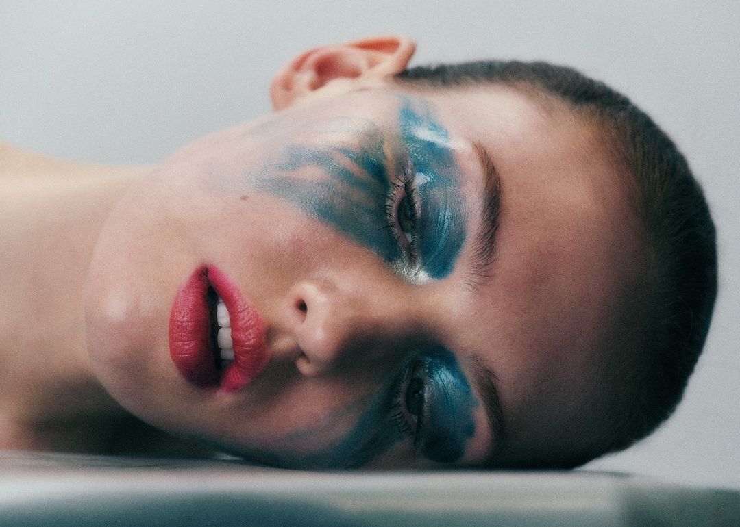 Woman with bold blue and white abstract makeup art and bright red lips laying against a light gray surface in a beauty shot.
