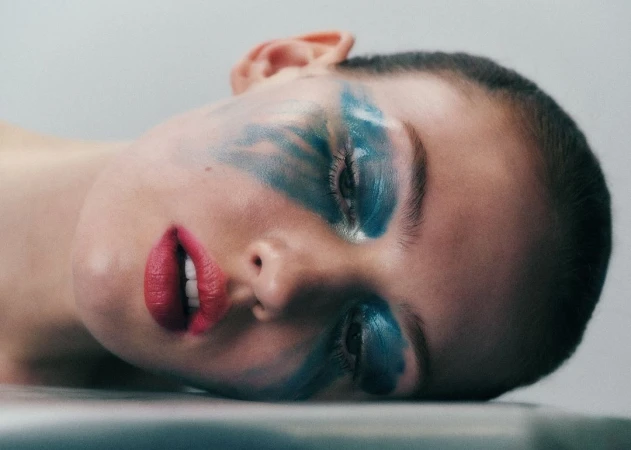 Close-up portrait of a woman with vibrant blue face paint and bright pink lipstick lying down on a neutral surface