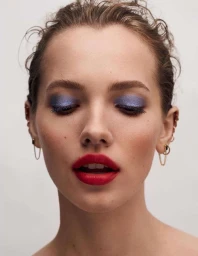 Close-up portrait of a woman with metallic blue eyeshadow and vibrant red lipstick, featuring delicate chain earrings.