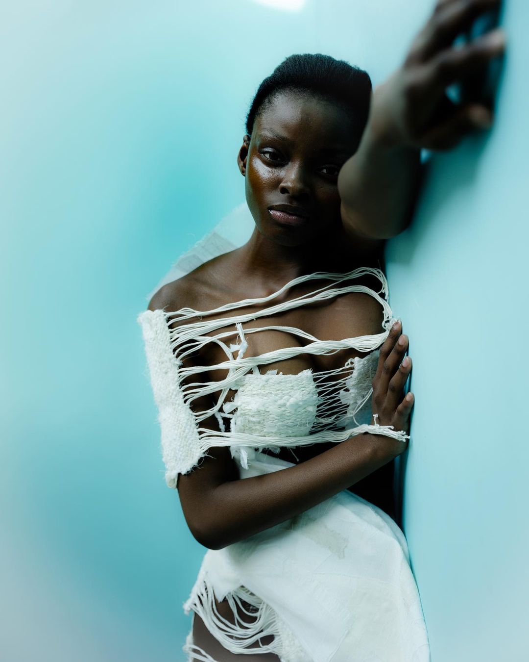 Striking portrait of a dark-skinned woman in a torn white top, with blue studio background and dramatic lighting accentuating her features.
