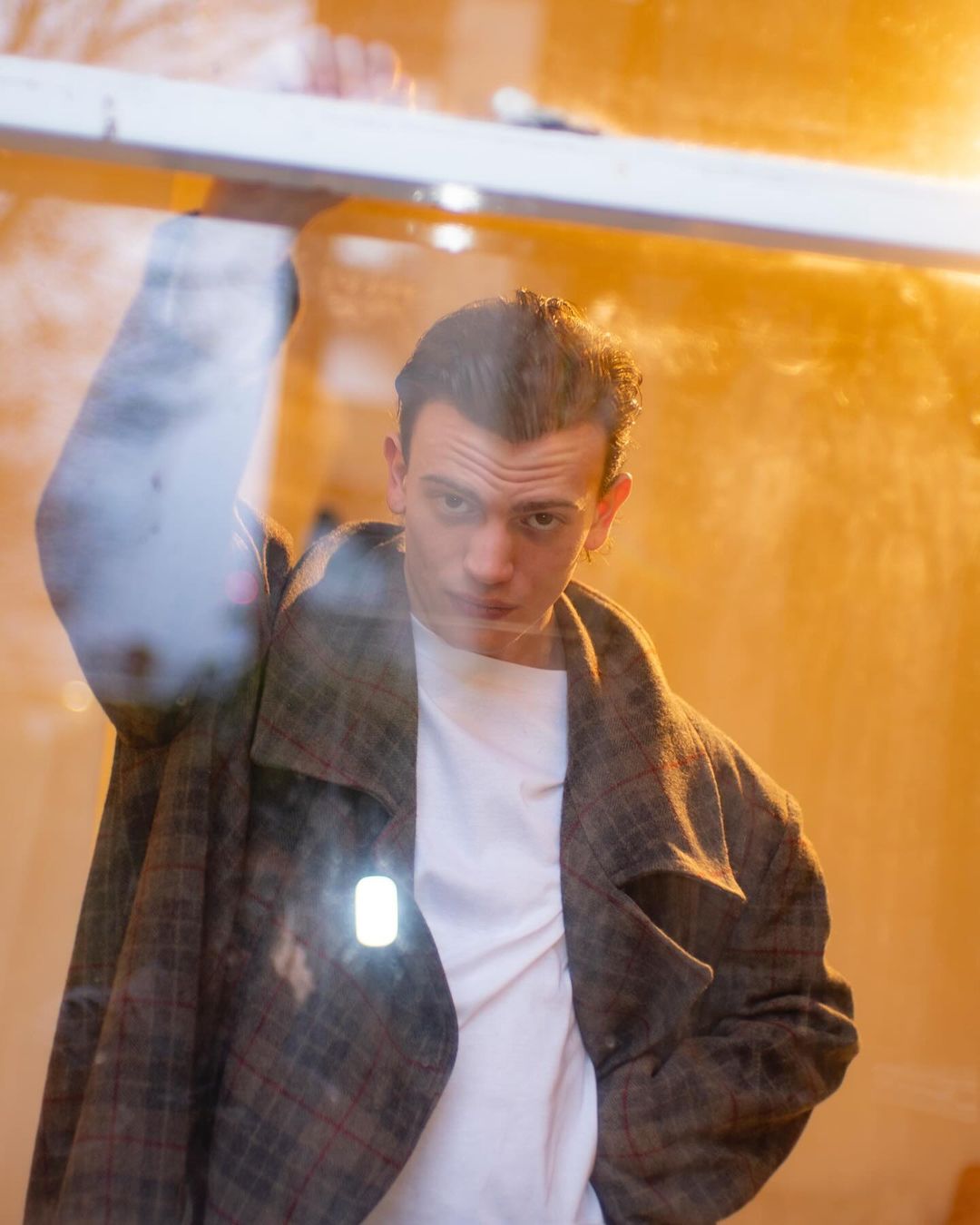Portrait of a young adult male wearing a plaid coat, standing indoors with soft lighting and a partially obscured background, shot at eye-level.