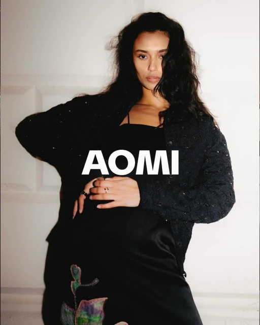 A young adult female model poses indoors wearing a black cardigan, black dress with a rose design, and rings on her fingers.