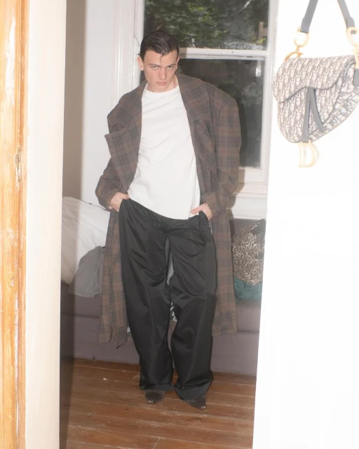 A slender man in a bathrobe and pajama pants stands in a doorway, looking at the camera with a handbag hanging on the door behind him.