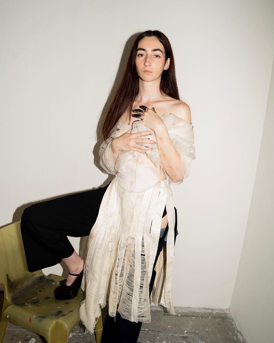 Young woman with long brown hair poses in a sheer, beige dress and black pants leaning against a yellow chair in a photo studio.