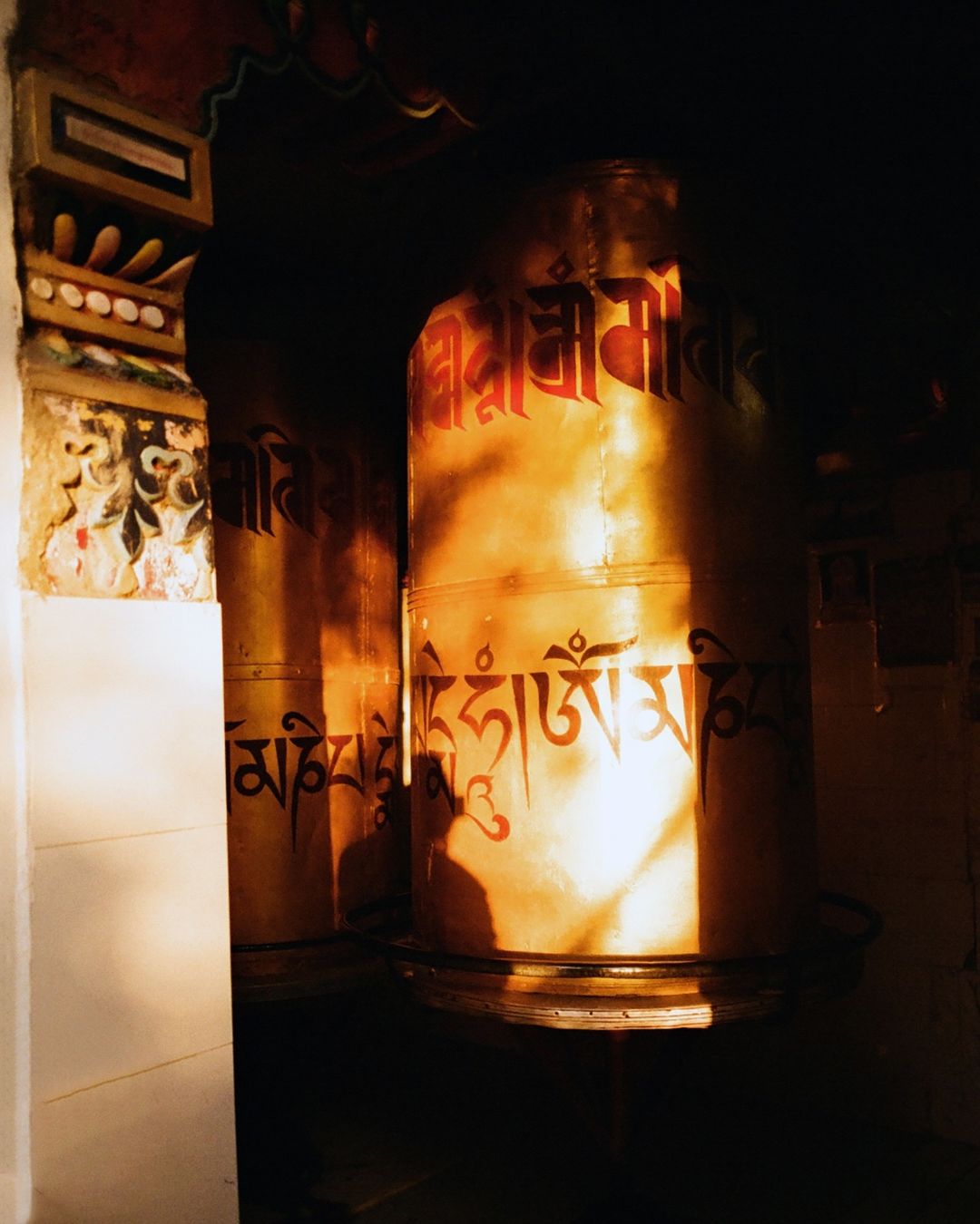 Illuminated prayer wheels adorned with Tibetan script, bathed in golden light, stand in a dimly lit interior space.