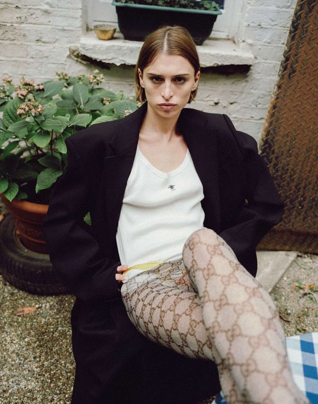 Model with short brown hair wearing an oversized black blazer over a white tank top and patterned tights, seated outdoors.