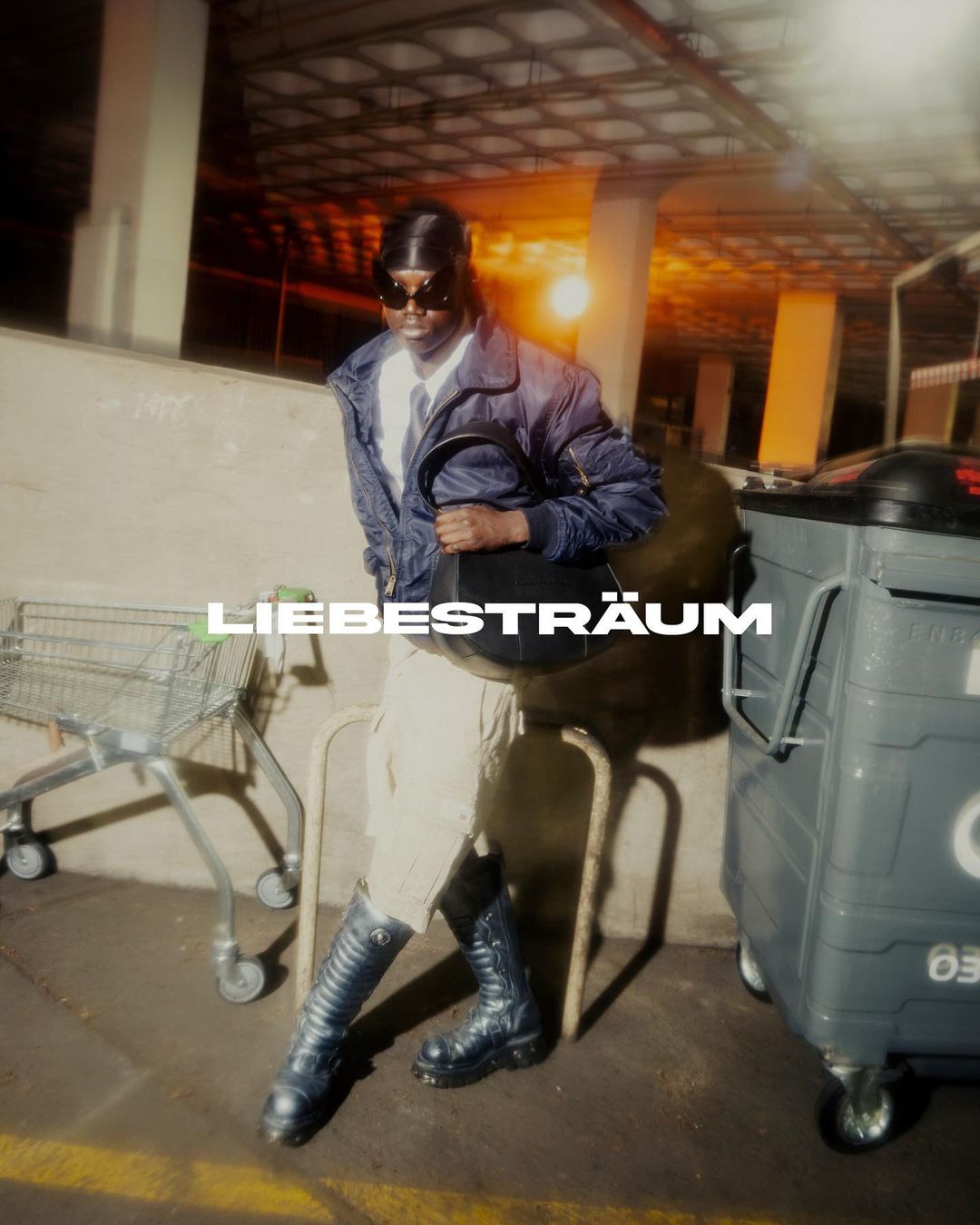 A person posing in an urban setting near a shopping cart and trash bin, wearing a jacket, boots and carrying a bag. 
