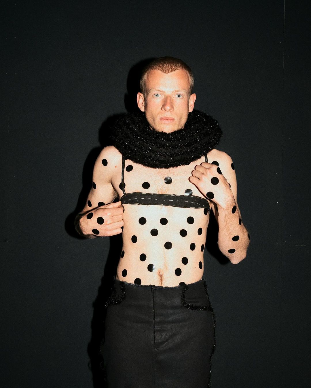 Man with polka dots on torso and arms wears black skirt and collar. Dark studio background. Fashion and style, posing.