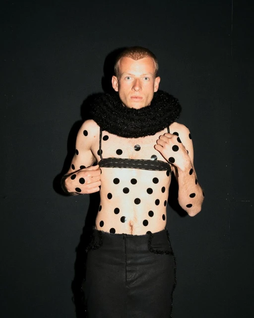 Man with polka dots on torso and arms wears black skirt and collar. Dark studio background. Fashion and style, posing.