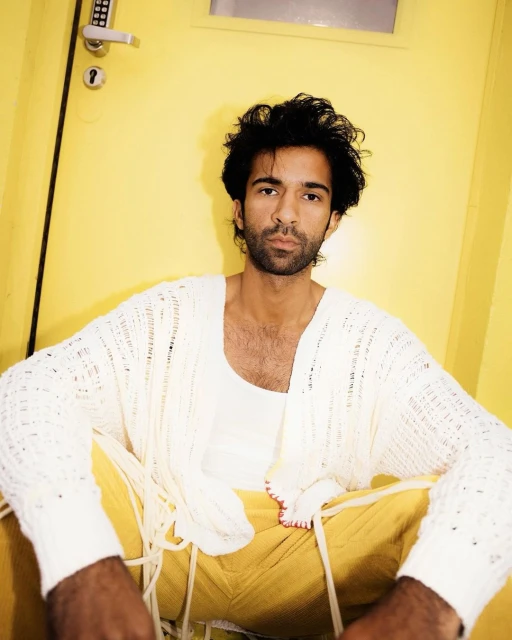 A stylish male model poses indoors against a yellow door, wearing a white cardigan, white tank top, and yellow pants in a studio setting.