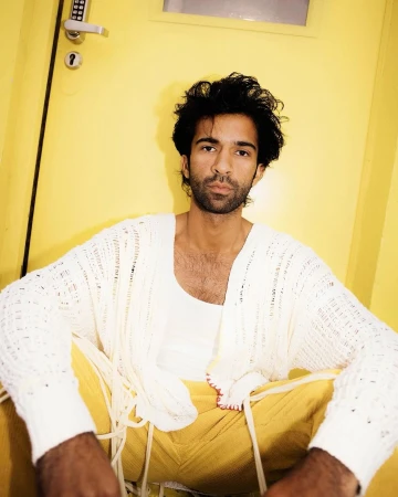 A stylish male model poses indoors against a yellow door, wearing a white cardigan, white tank top, and yellow pants in a studio setting.