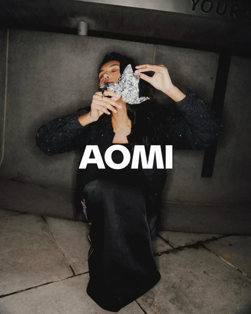 Person holding silver foil object in a dark setting, kneeling, with a textured wall background, and large AOMI text overlay.