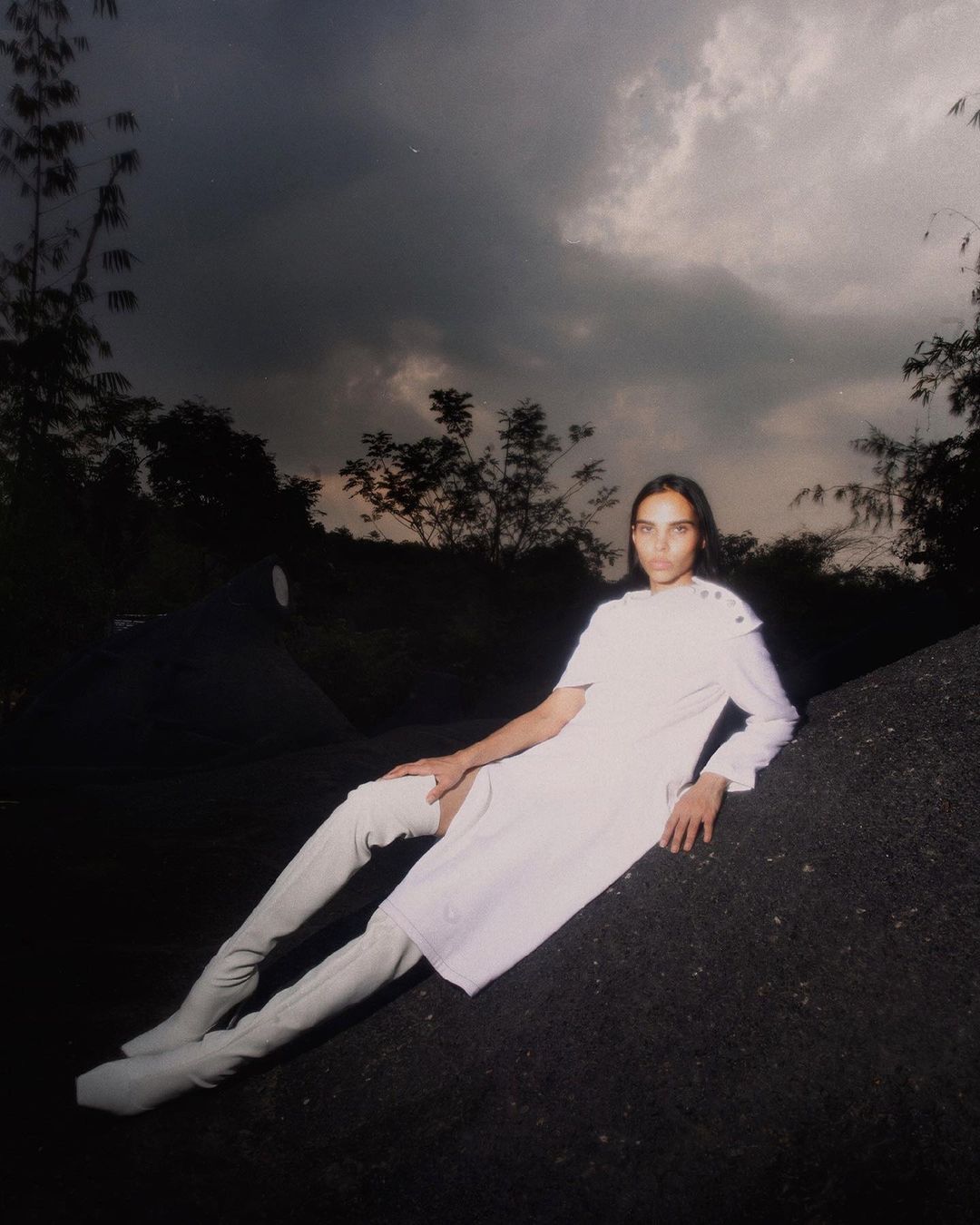 Woman in a white dress and thigh-high boots reclines on a rock under a cloudy sky in an outdoor fashion shot with nature background.