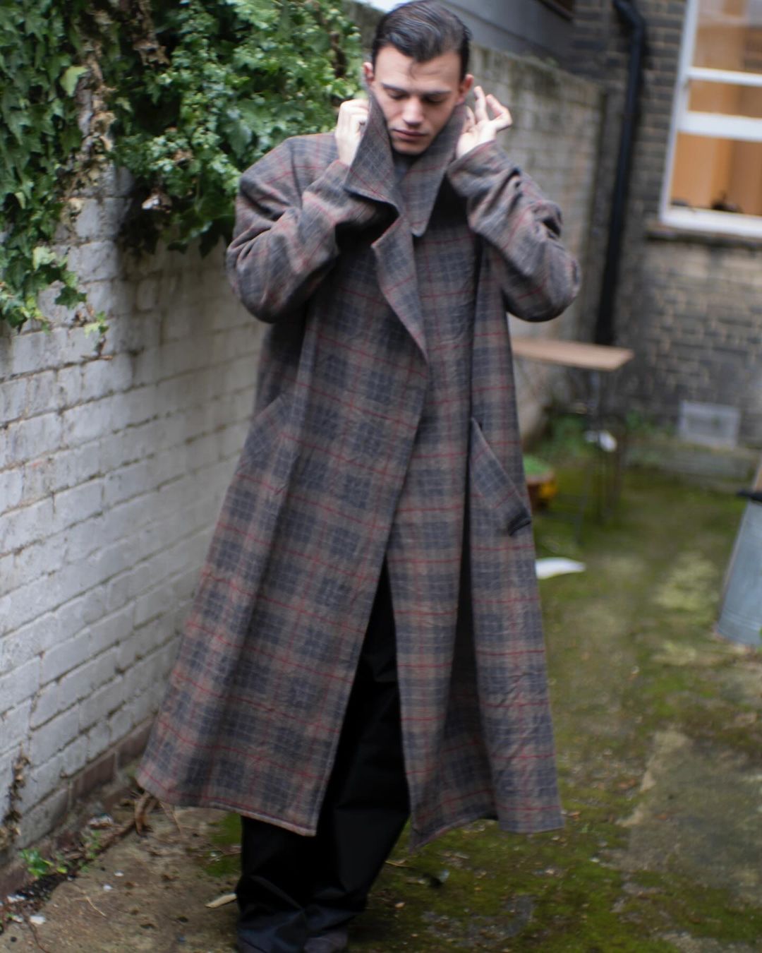 Man wearing a long plaid coat outdoors in an alleyway, adjusting the high collar with his hands.