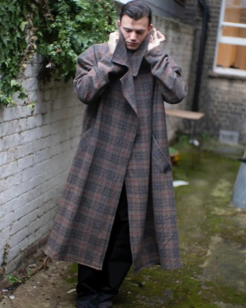 Man wearing a long plaid coat outdoors in an alleyway, adjusting the high collar with his hands.