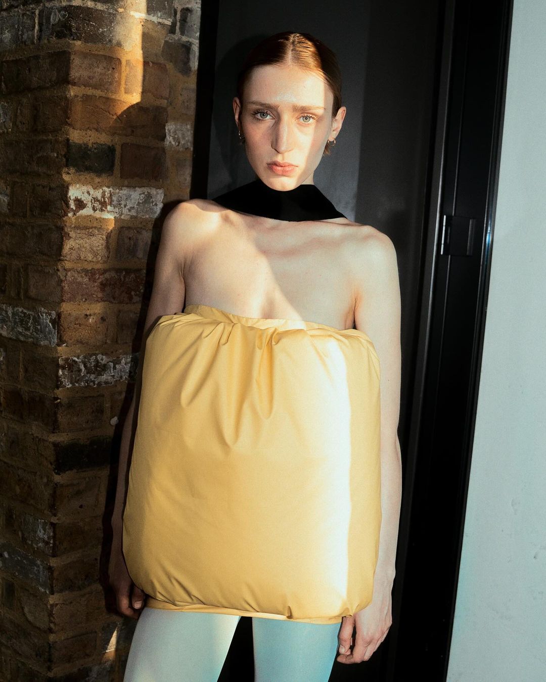 A woman in a yellow garment stands in front of a brick wall, bathed in sunlight, casting shadows on the weathered surface.