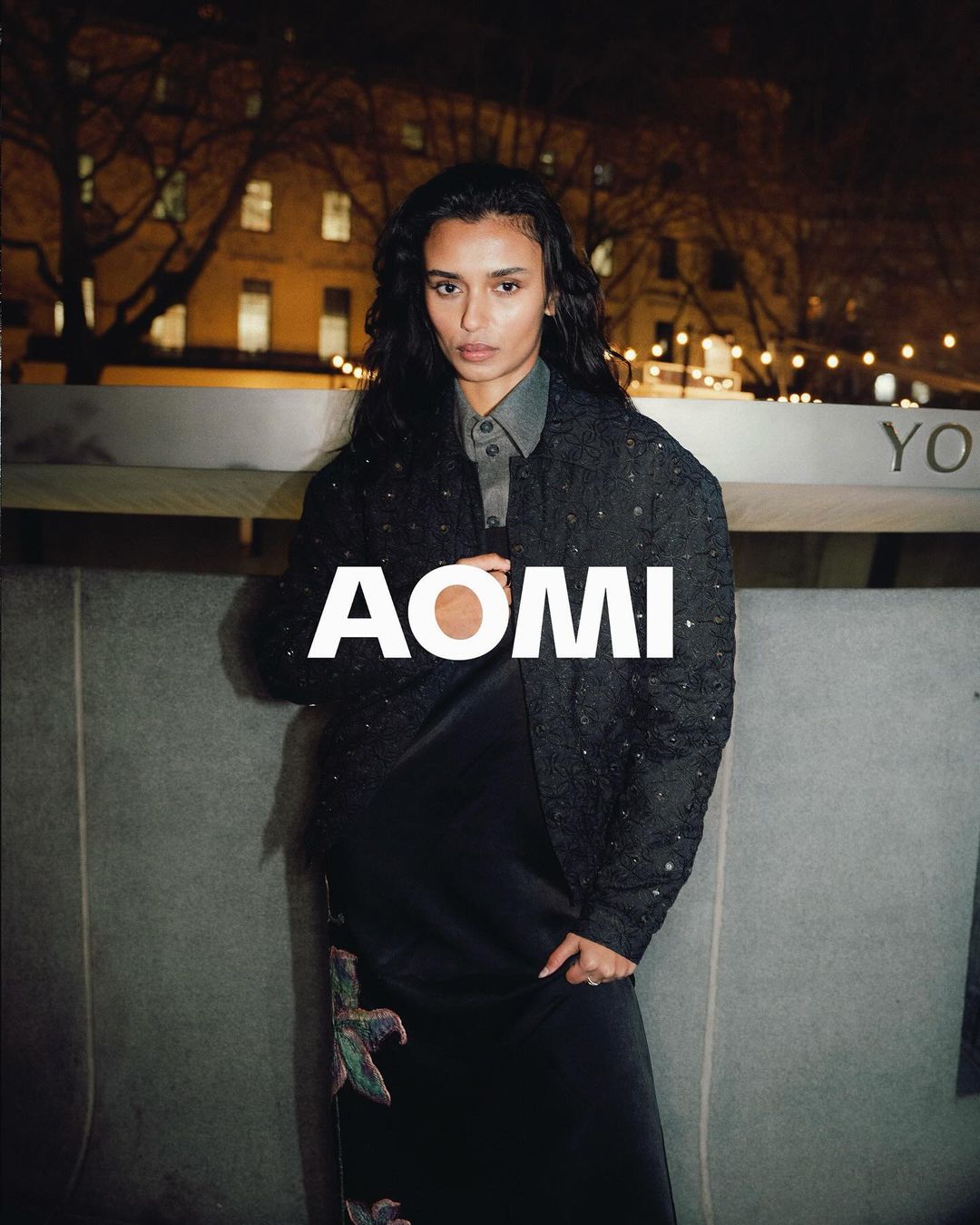 Adult woman with long dark hair poses in a grey silk dress and patterned jacket in an outdoor urban environment at night.