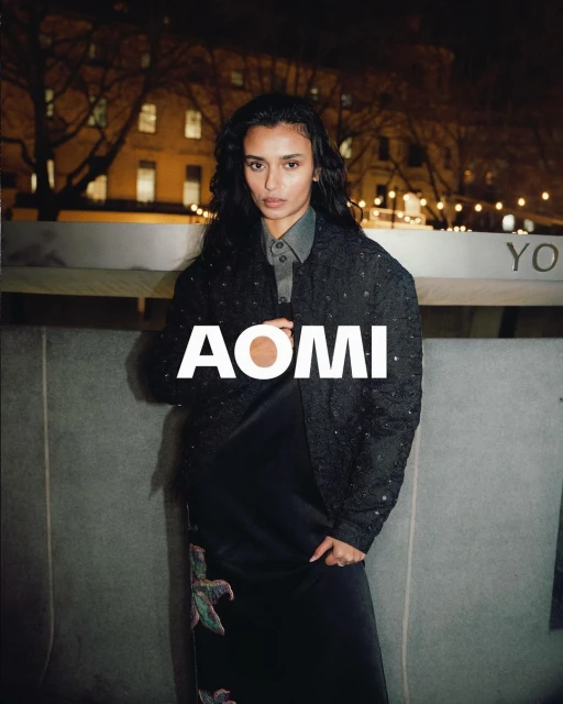 Adult woman with long dark hair poses in a grey silk dress and patterned jacket in an outdoor urban environment at night.