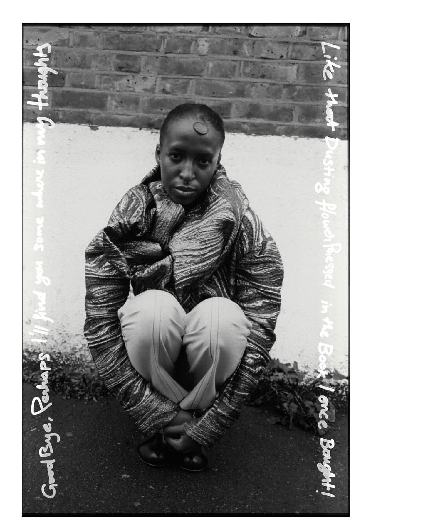 Monochrome portrait of a person crouching with a serious expression, wearing patterned jacket and pale pants against a brick wall.