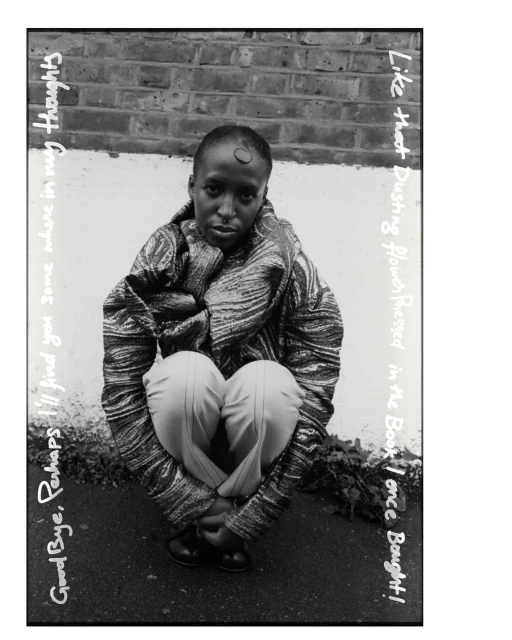 Monochrome portrait of a person crouching with a serious expression, wearing patterned jacket and pale pants against a brick wall.