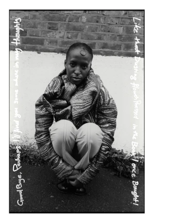 Monochrome portrait of a person crouching with a serious expression, wearing patterned jacket and pale pants against a brick wall.