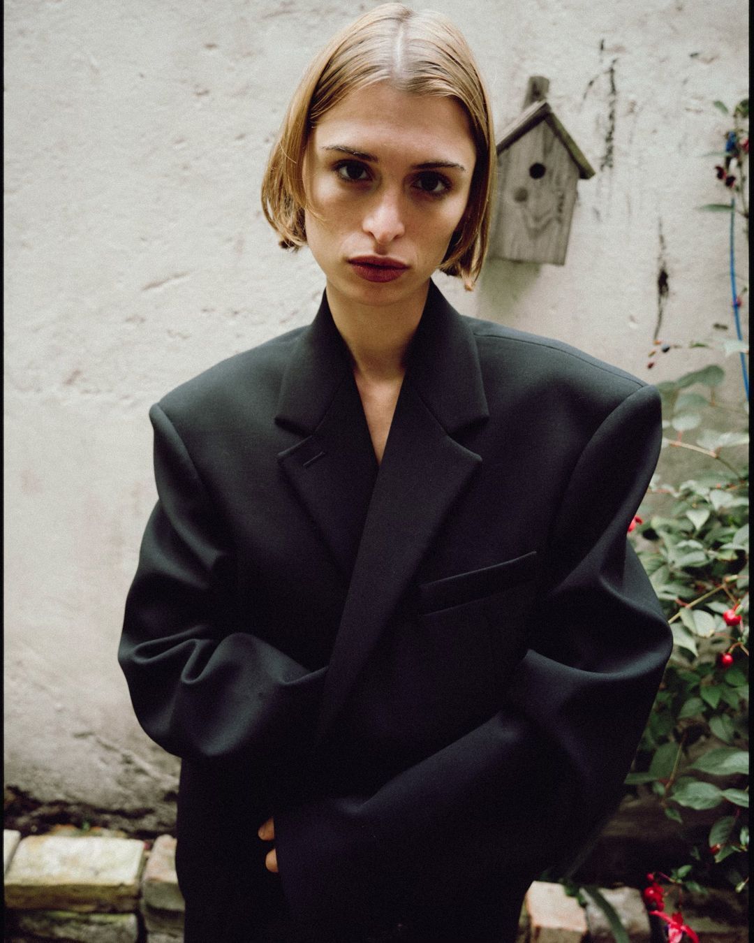 Blonde woman in an oversized black blazer poses in front of a weathered wall with a birdhouse and greenery in a natural outdoor setting.
