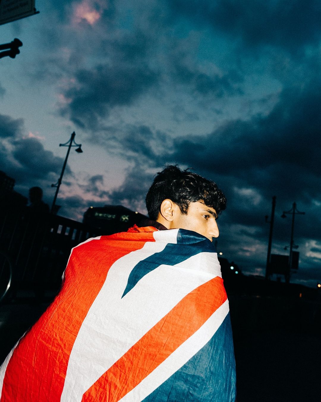Adult male draped in a Union Jack flag stands outside at dusk with a cloudy sky in the background; low-angle shot.