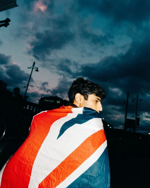 Adult male draped in a Union Jack flag stands outside at dusk with a cloudy sky in the background; low-angle shot.