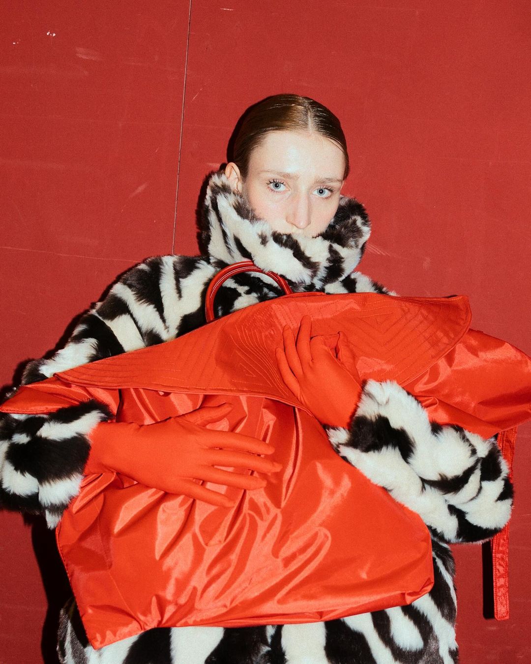 Fashion portrait of a fair-skinned woman wearing a zebra-print faux fur coat, vibrant red gloves, and a matching oversized bag against a red wall.