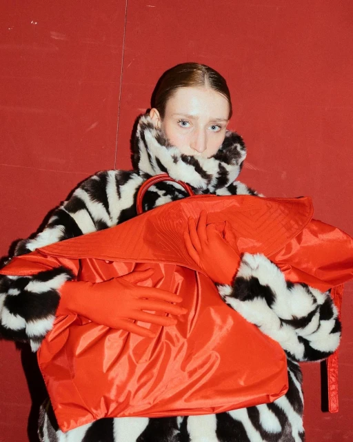 Fashion portrait of a fair-skinned woman wearing a zebra-print faux fur coat, vibrant red gloves, and a matching oversized bag against a red wall.