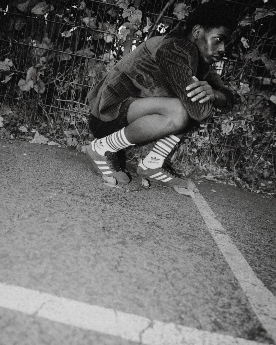 Monochrome outdoor portrait of a young Black male crouching, wearing Adidas sneakers and socks, with greenery and a fence in the background.