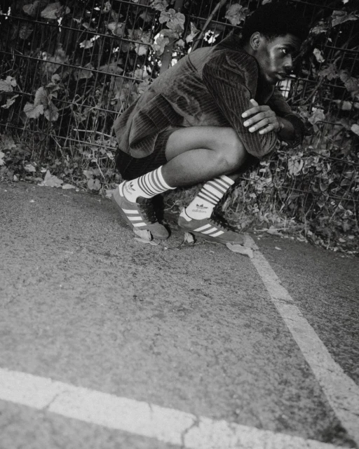 Monochrome outdoor portrait of a young Black male crouching, wearing Adidas sneakers and socks, with greenery and a fence in the background.