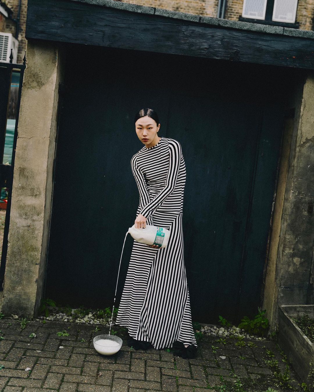 Asian woman in a striped dress pours milk into a bowl outdoors. Editorial fashion shot in an urban setting with natural lighting.