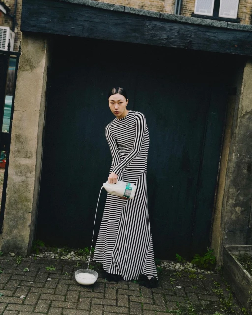 Asian woman in a striped dress pours milk into a bowl outdoors. Editorial fashion shot in an urban setting with natural lighting.