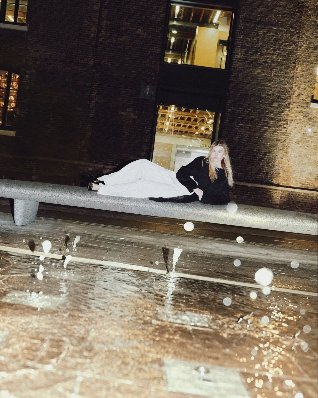 Blonde woman in black shirt and white pants laying on concrete bench, brick building and water fountain in background at night.