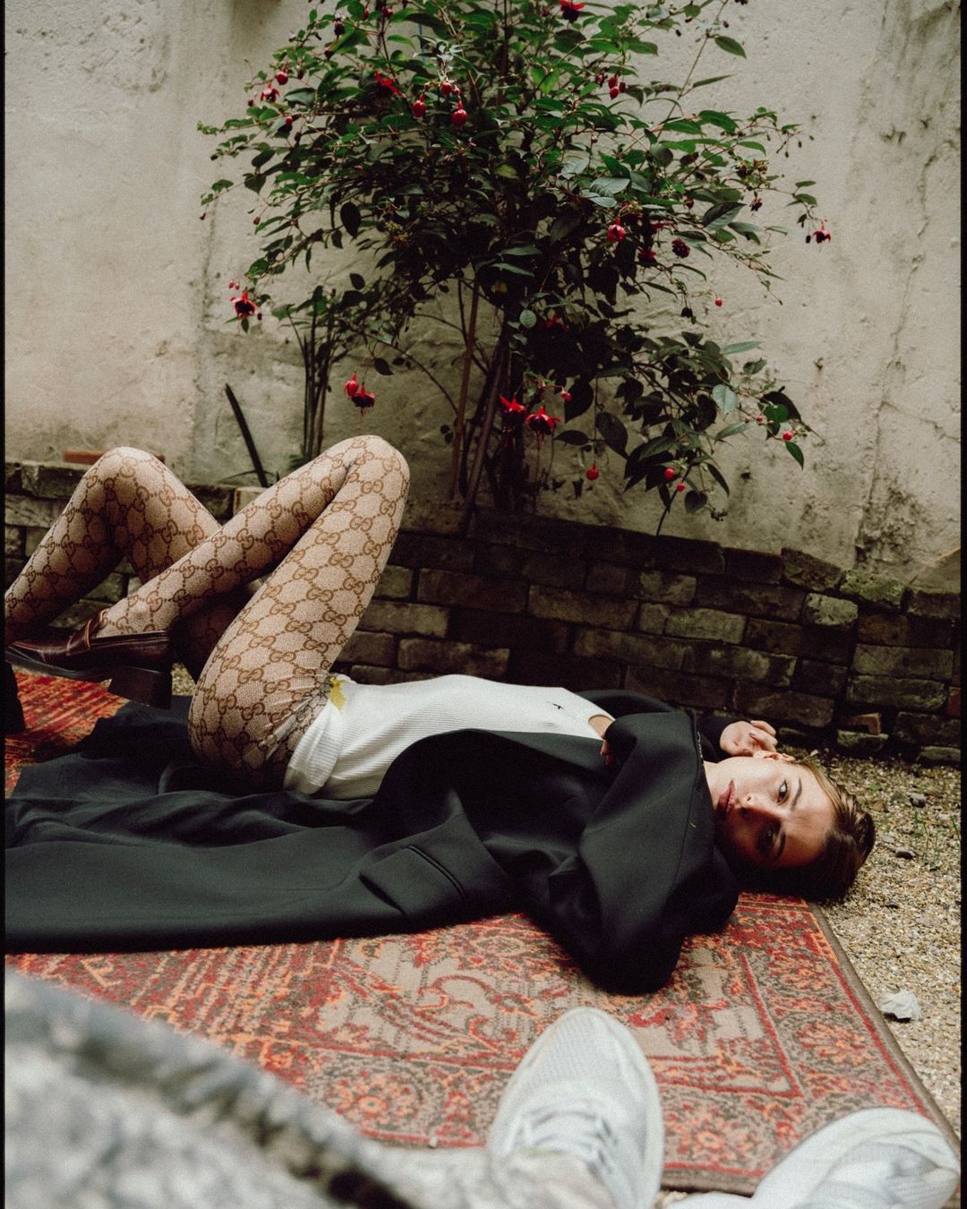 Woman in patterned tights lies on an oriental rug in a walled courtyard, with a flowering bush, wearing a white shirt and black jacket.