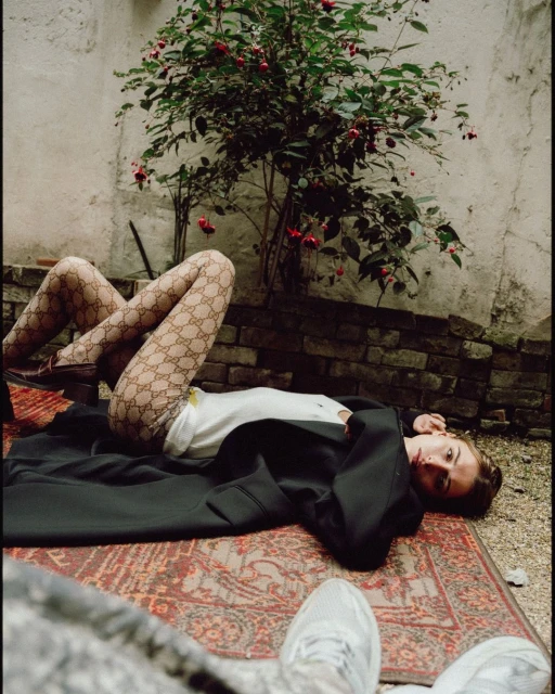 Woman in patterned tights lies on an oriental rug in a walled courtyard, with a flowering bush, wearing a white shirt and black jacket.