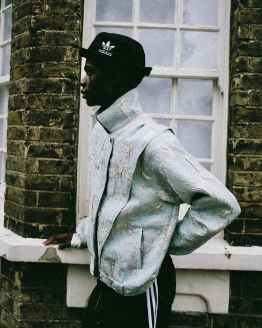 An androgynous person with dark skin in an pale blue jacket and black Adidas hat poses against a brick wall and window frame outside.