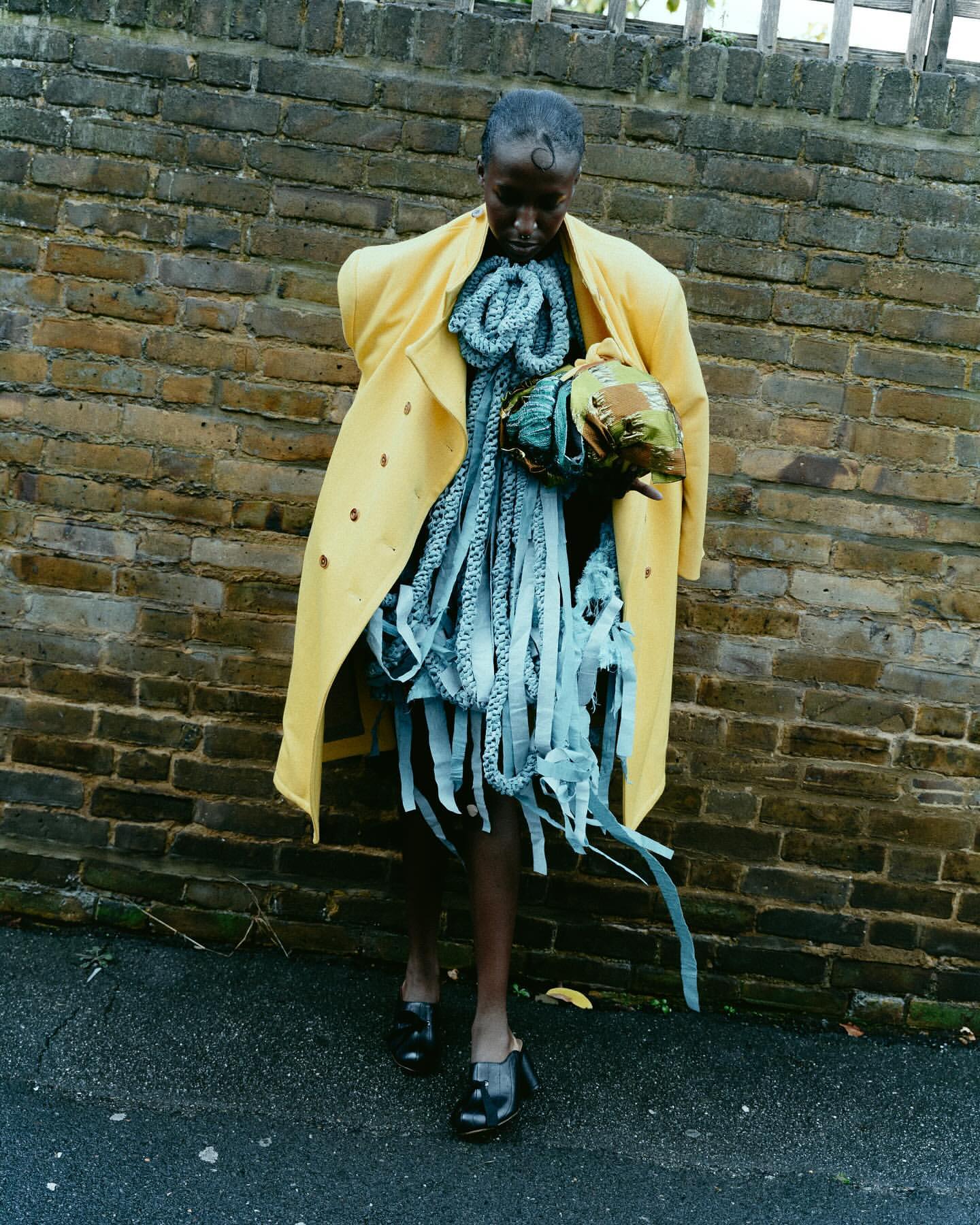 Fashionable Black woman in yellow coat and avant-garde blue dress poses against a brick wall, showcasing a unique style with dark shoes.