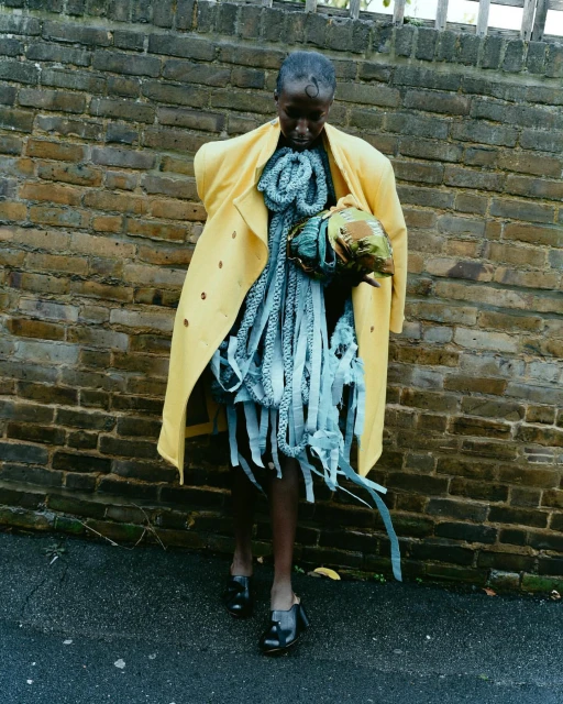 Fashionable Black woman in yellow coat and avant-garde blue dress poses against a brick wall, showcasing a unique style with dark shoes.