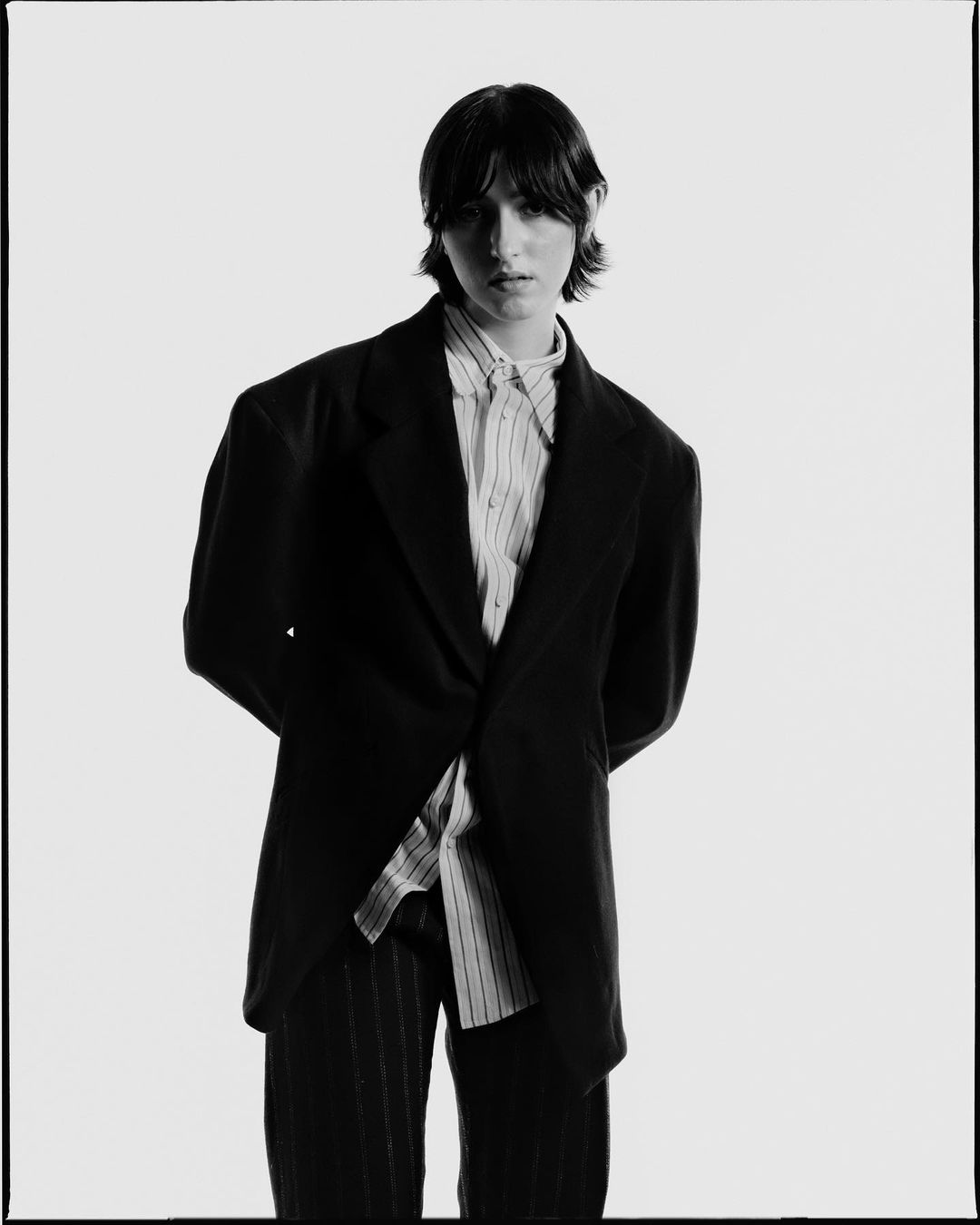 A young adult poses in a black blazer and striped shirt and pants against a white background, delivering a high-fashion look.