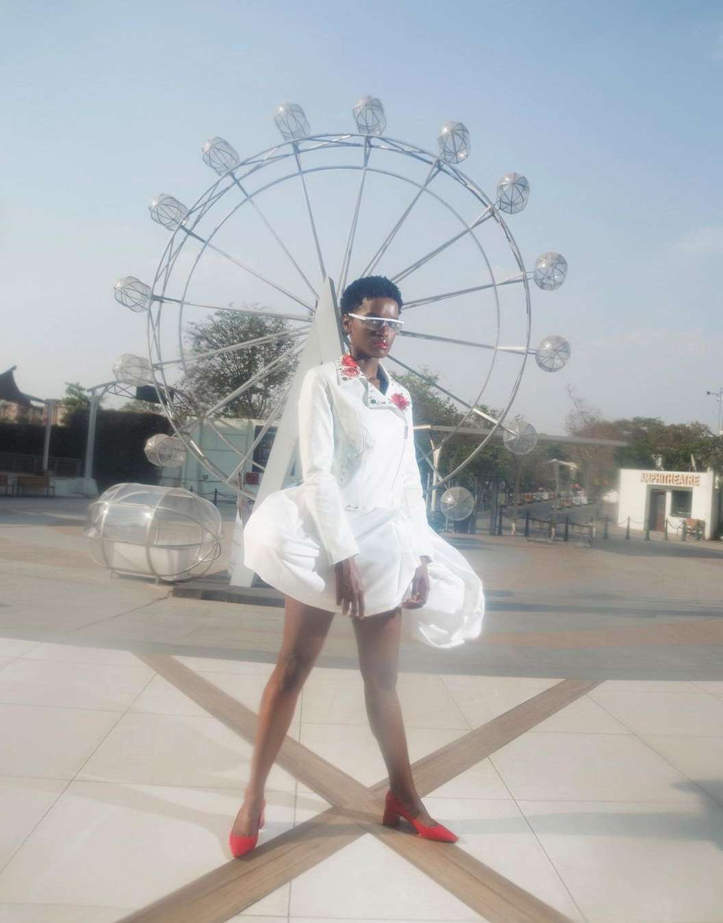 A fashionably dressed person poses in front of an outdoor metal sculpture. The person wears a white dress and red shoes in daylight.
