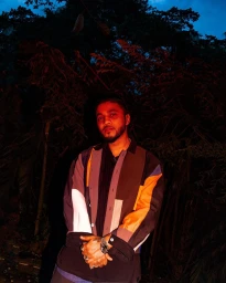 Portrait of a man wearing a color-blocked patterned shirt and rings, lit with red light against a dark foliage background.