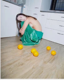 Woman in green velvet skirt crouching on wooden floor in kitchen with scattered oranges