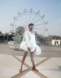 Model in white structured jacket and flared mini skirt wearing red shoes standing before a large Ferris wheel.