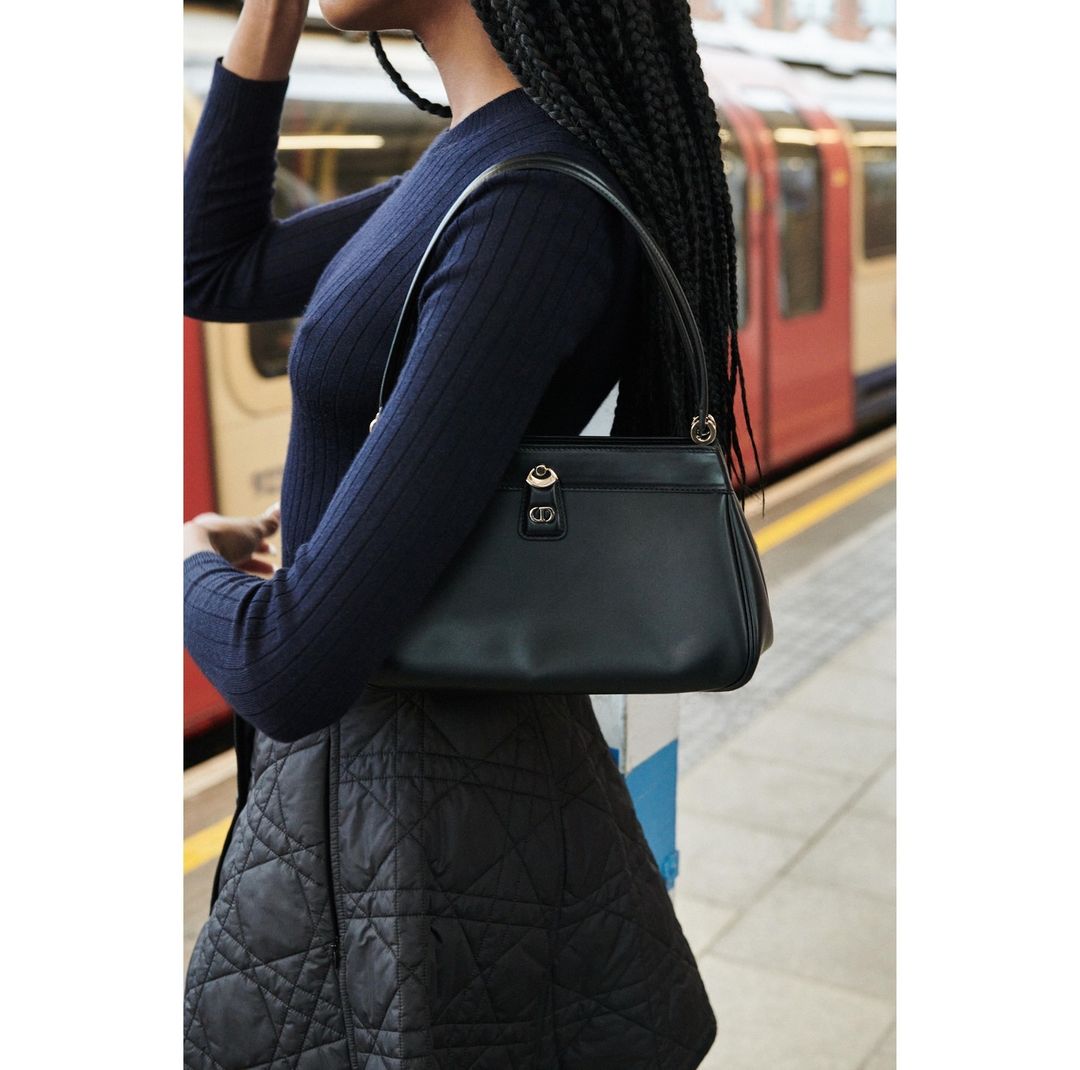 Woman in a navy sweater and skirt stands on a train platform with a black leather handbag in front of a red train.